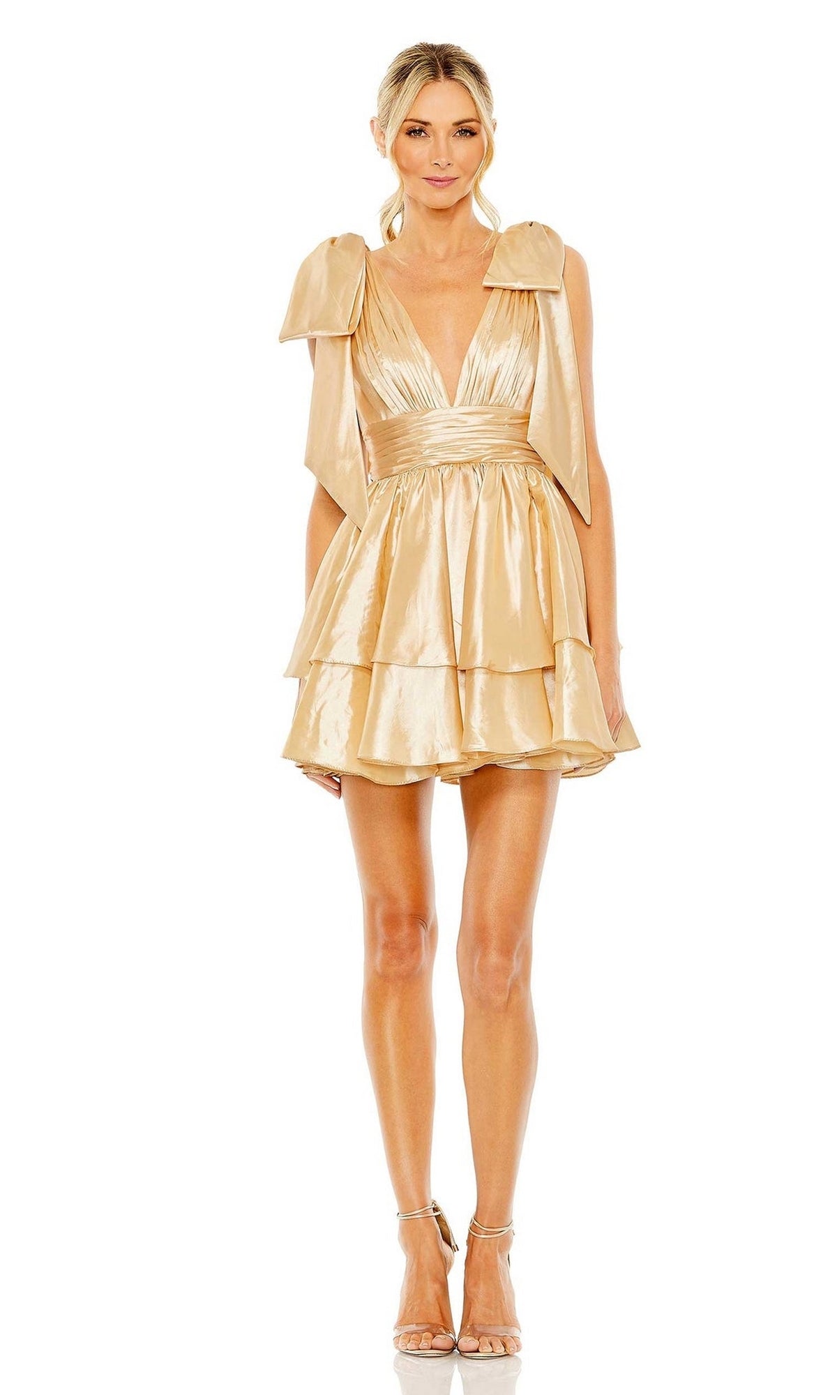 Woman models the Mac Duggal 11657 Bow Short Ruffled Hoco Dress in shiny gold, featuring a deep V-neckline and shoulder bows, paired with strappy heels against a white background.
